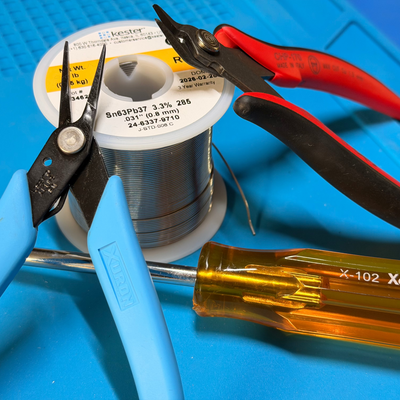 Needle-nosed pliers, solder, wire cutters, and a screwdriver on a blue mat. 